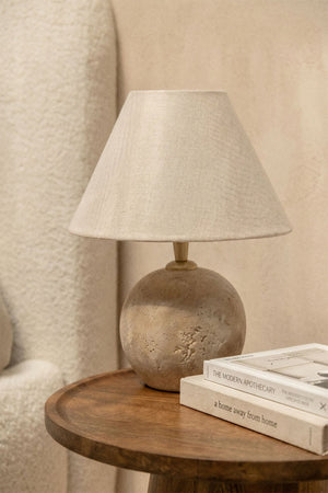 Marvexi table lamp – full studio shot on warm beige background, round travertine stone base with E27 holder and natural linen empire shade, cable visible