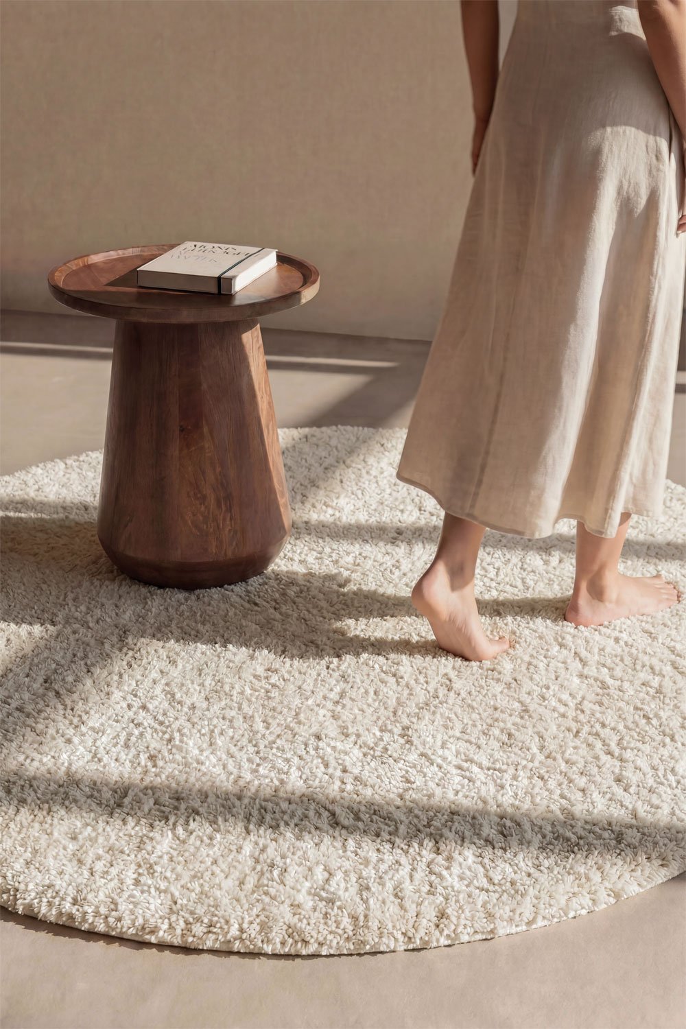 Daroca round rug Ø120 cm in cream beige – lifestyle shot with walnut side table and person in linen trousers standing on rug